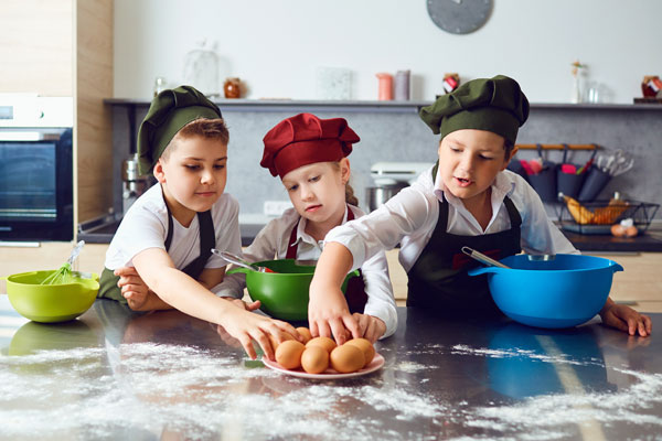 Zdjęcie przedstawia trójkę dzieci przygotowującą potrawę na stole kuchennym